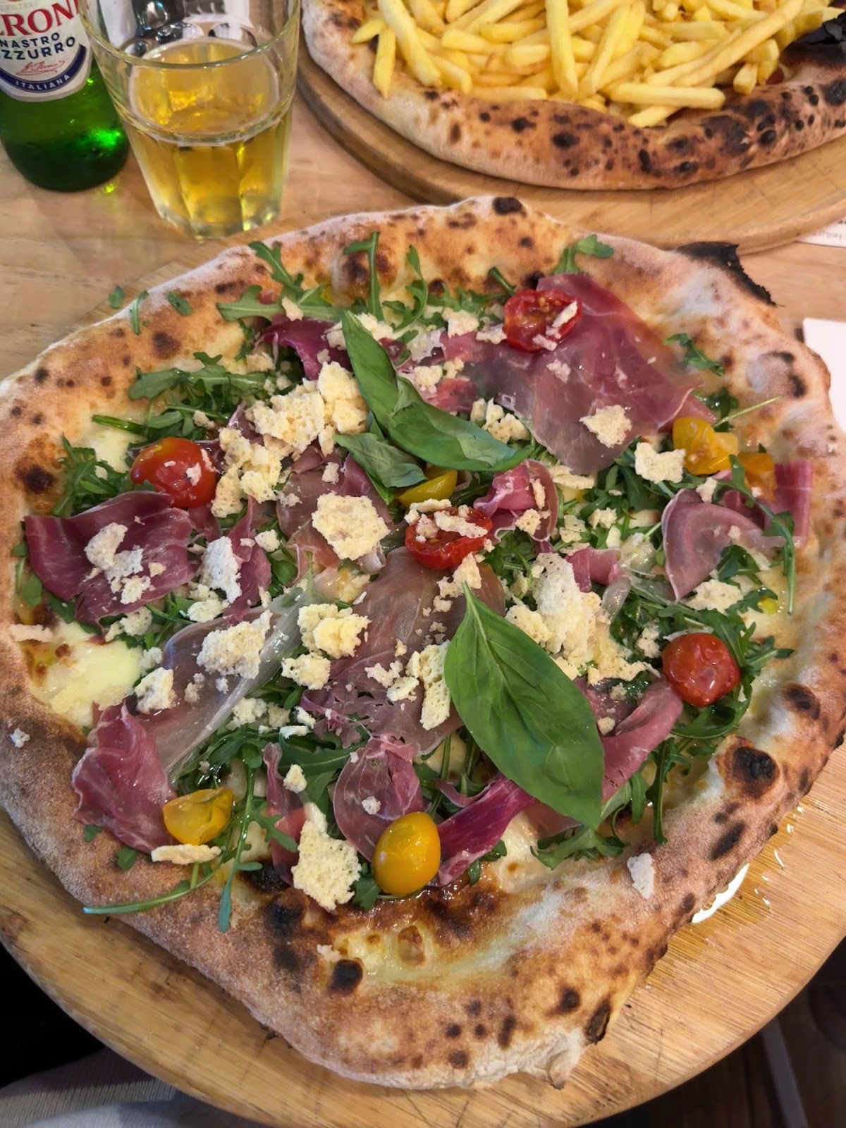 Pizza topped with rocket, cured meat and shaved cheese on a wooden board, beer and fries behind