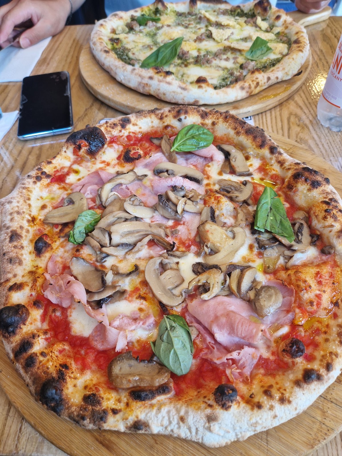 Ham and mushroom pizza in the foreground with a second pizza behind on a busy table